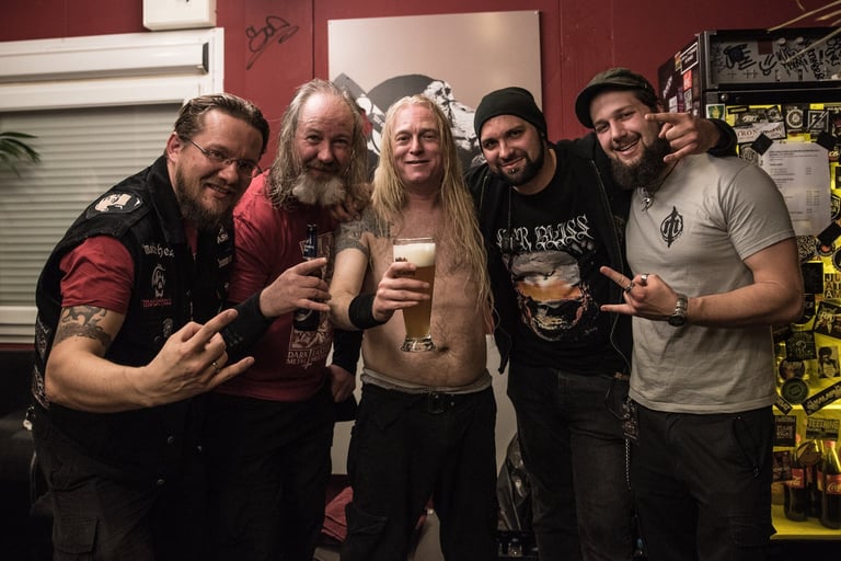 Fünf Männer mit Bärten posieren zusammen in einem dekorierten Raum, halten Getränke, tragen Casual- und Band-T-Shirts